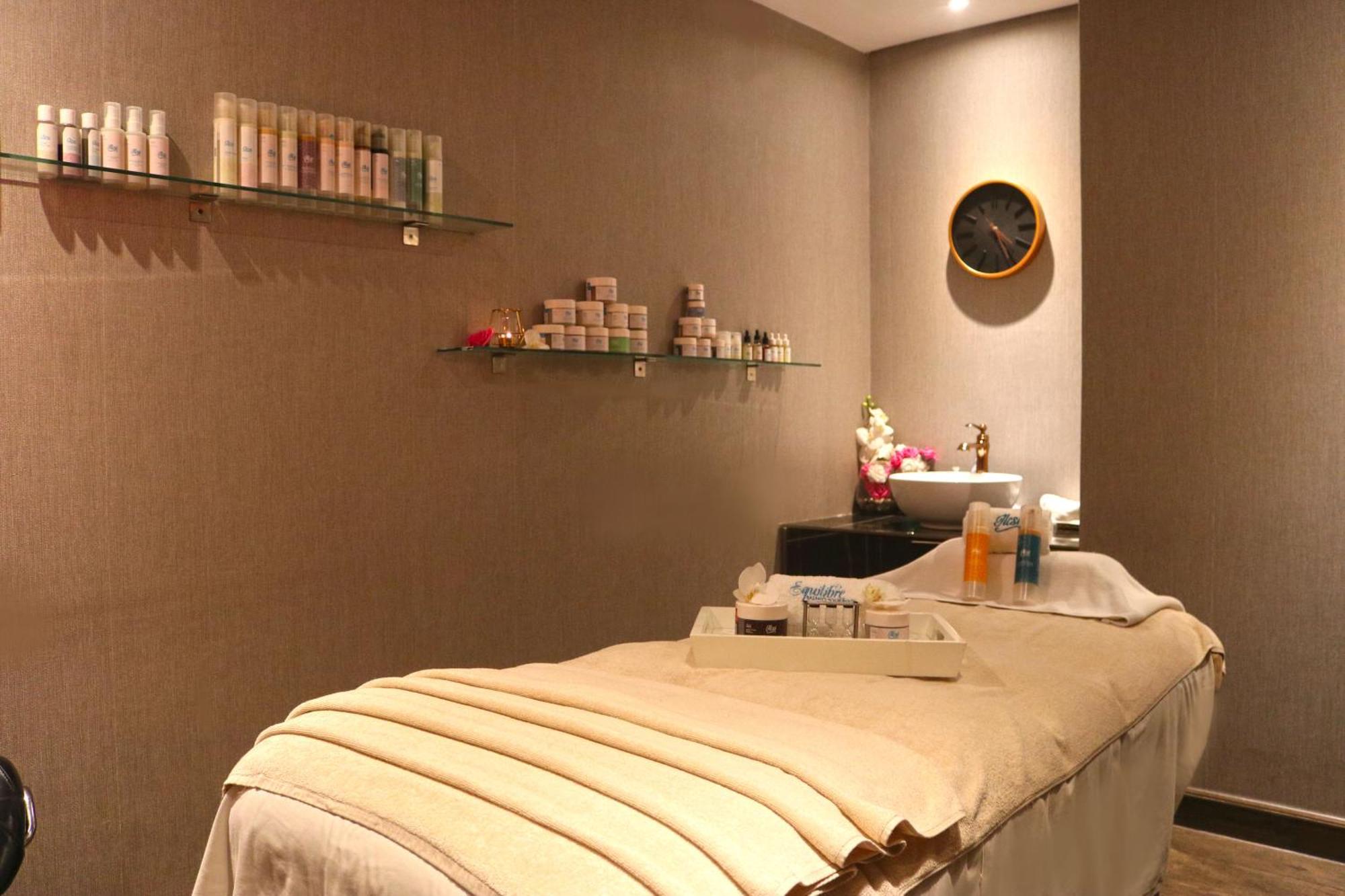 Lagoona Beach Luxury Resort And Spa Манама Экстерьер фото The photo shows a spa treatment room. There is a massage table draped with a light-colored blanket, creating a relaxing atmosphere. On the wall, there are shelves displaying various skincare products in bottles and jars. A clock is mounted on the wal