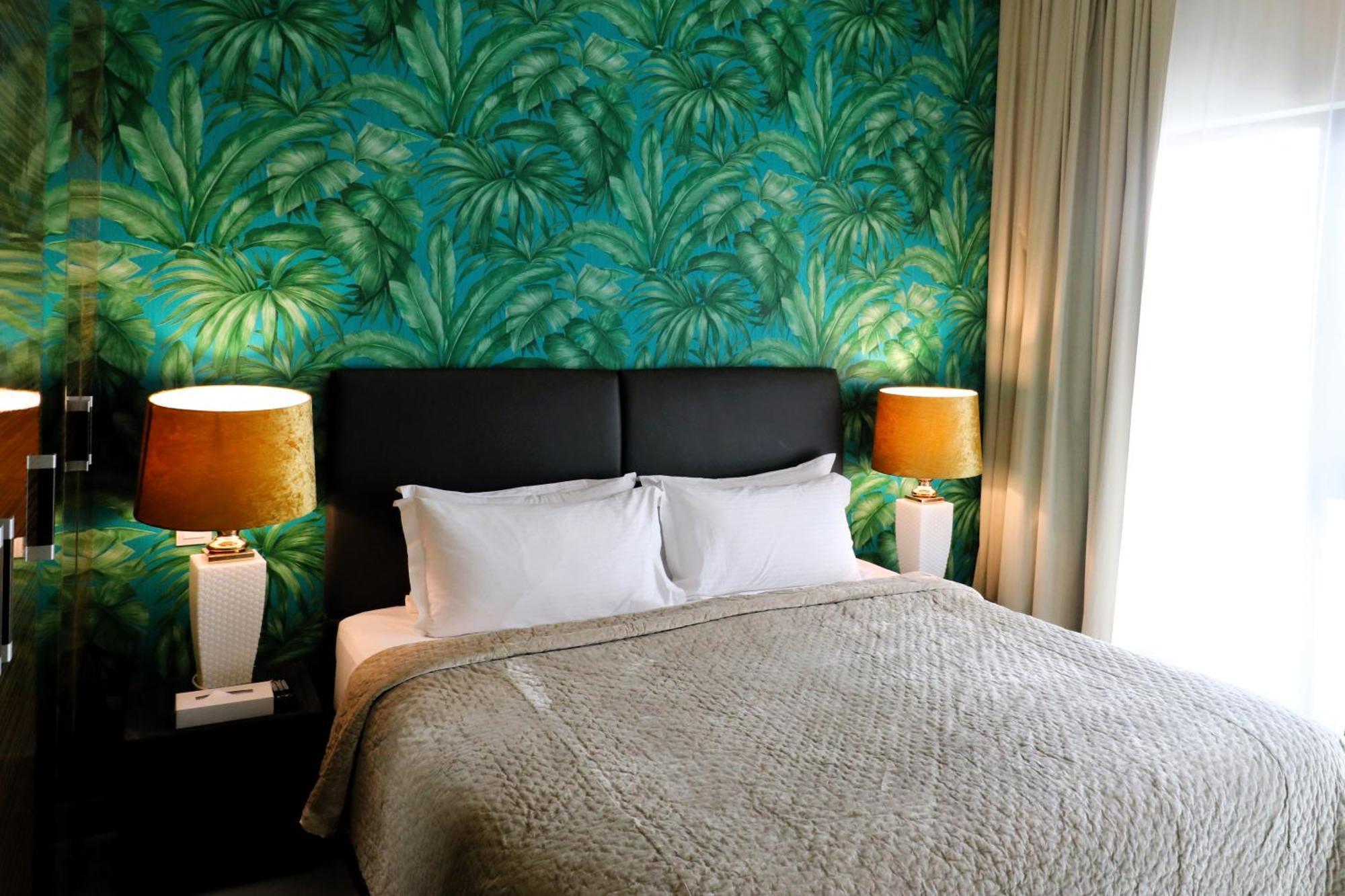 Lagoona Beach Luxury Resort And Spa Манама Экстерьер фото The photo shows a stylish bedroom featuring a large bed with white pillows and a gray blanket. The headboard is upholstered in black, providing a modern touch. Behind the bed, there is a vibrant green wallpaper with a tropical leaf pattern, adding a 